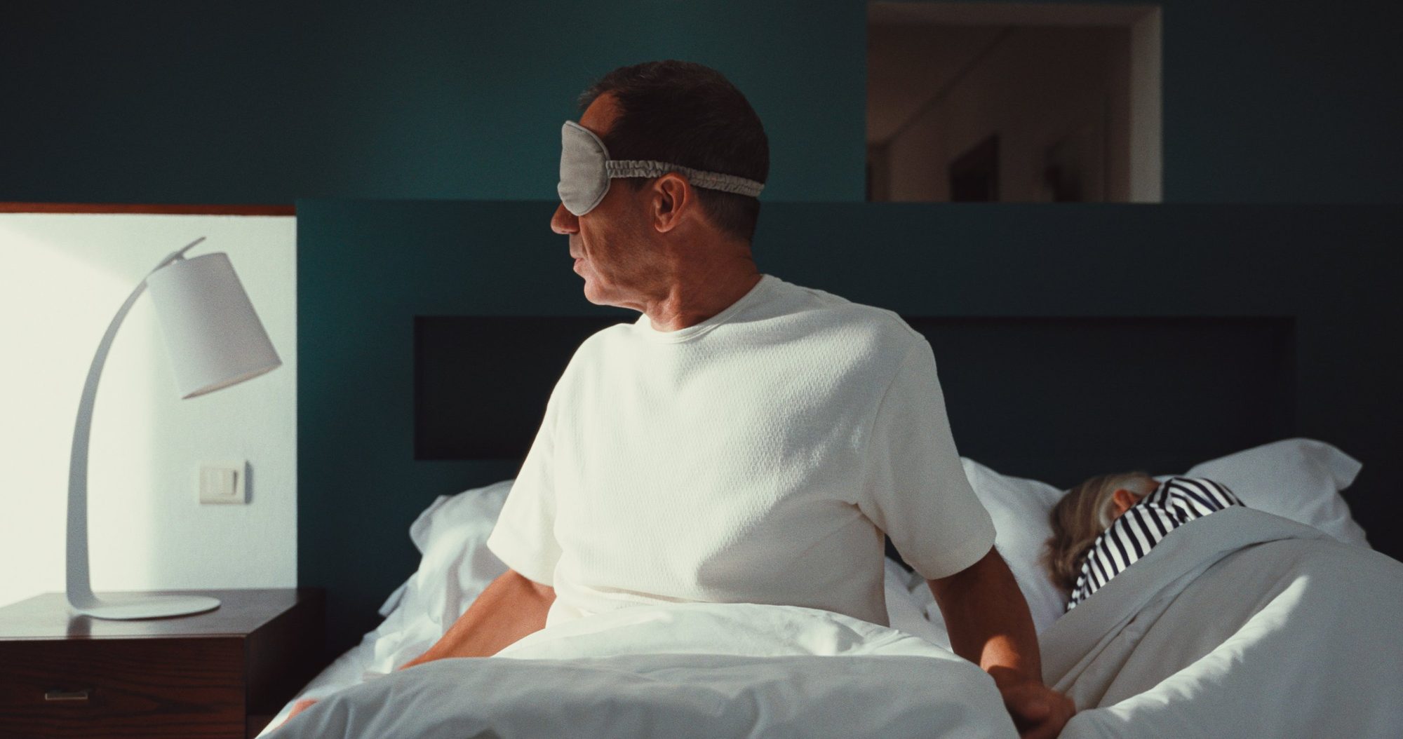 A man in a sleep mask sits up in a teal hotel room