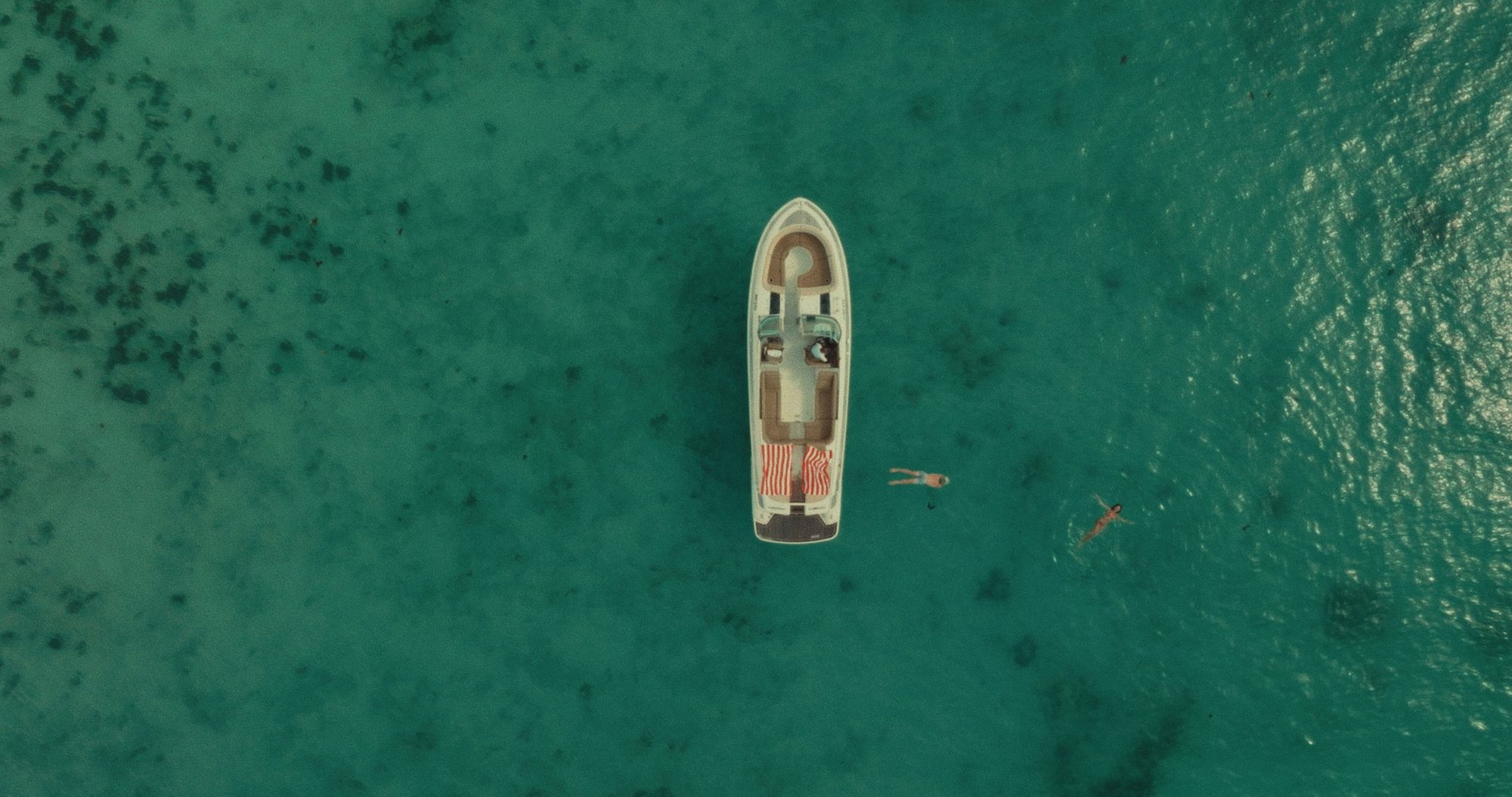 Overhead: a white boat on turquoise water with two swimmers beside it