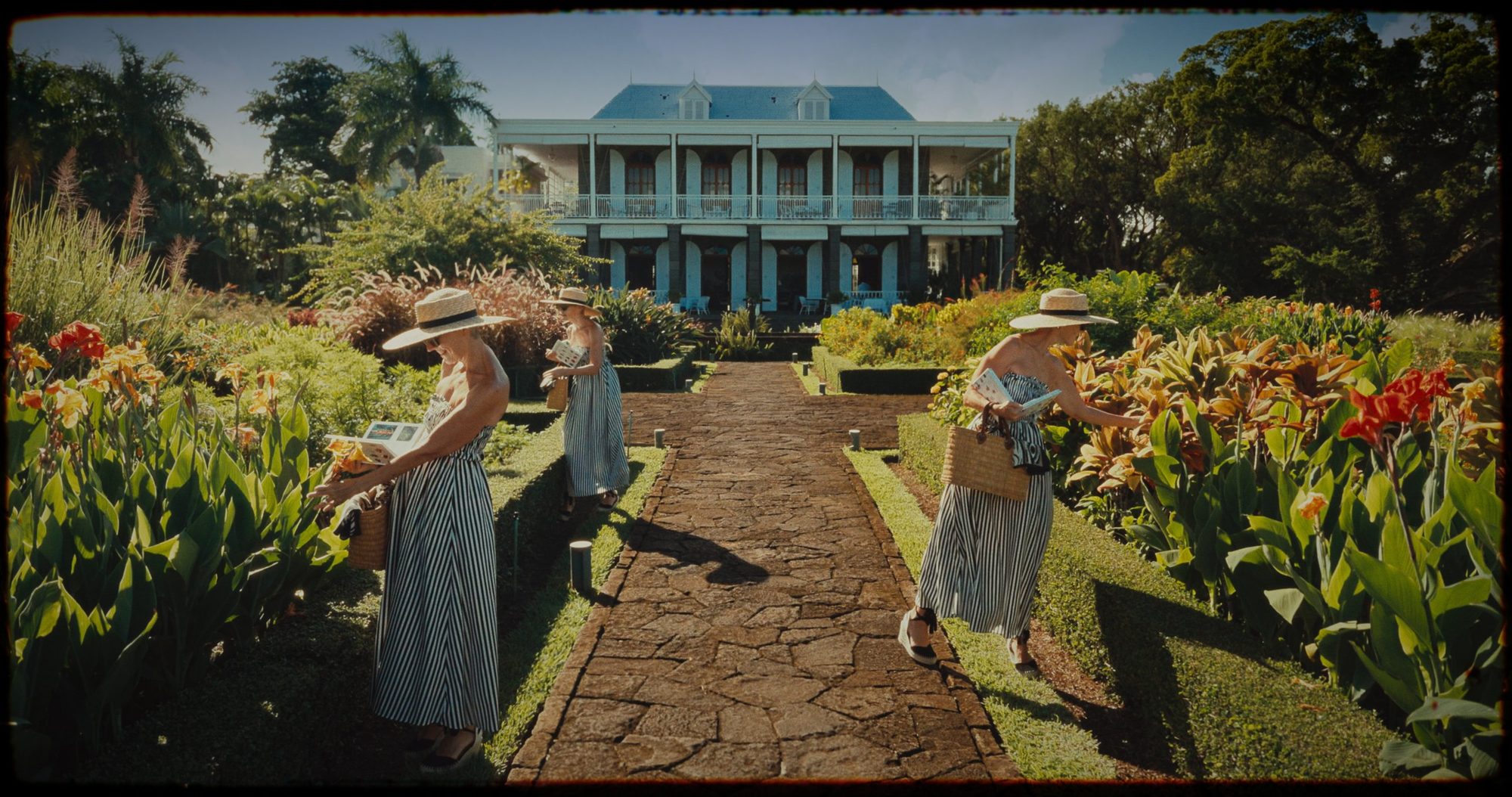 A woman in a striped dress appears three times along a garden path to a colonial house
