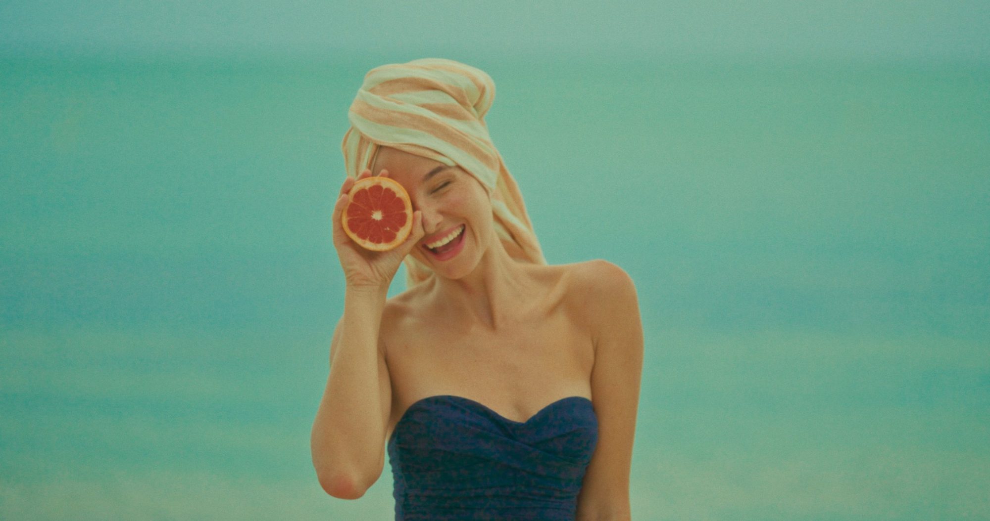 A laughing woman in a towel turban holds a grapefruit to her eye