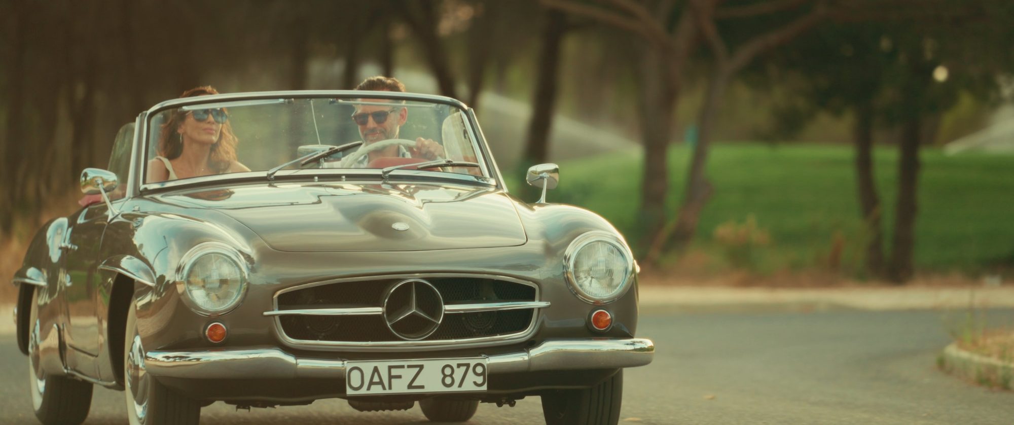 A couple drives a silver vintage Mercedes down a tree-lined road
