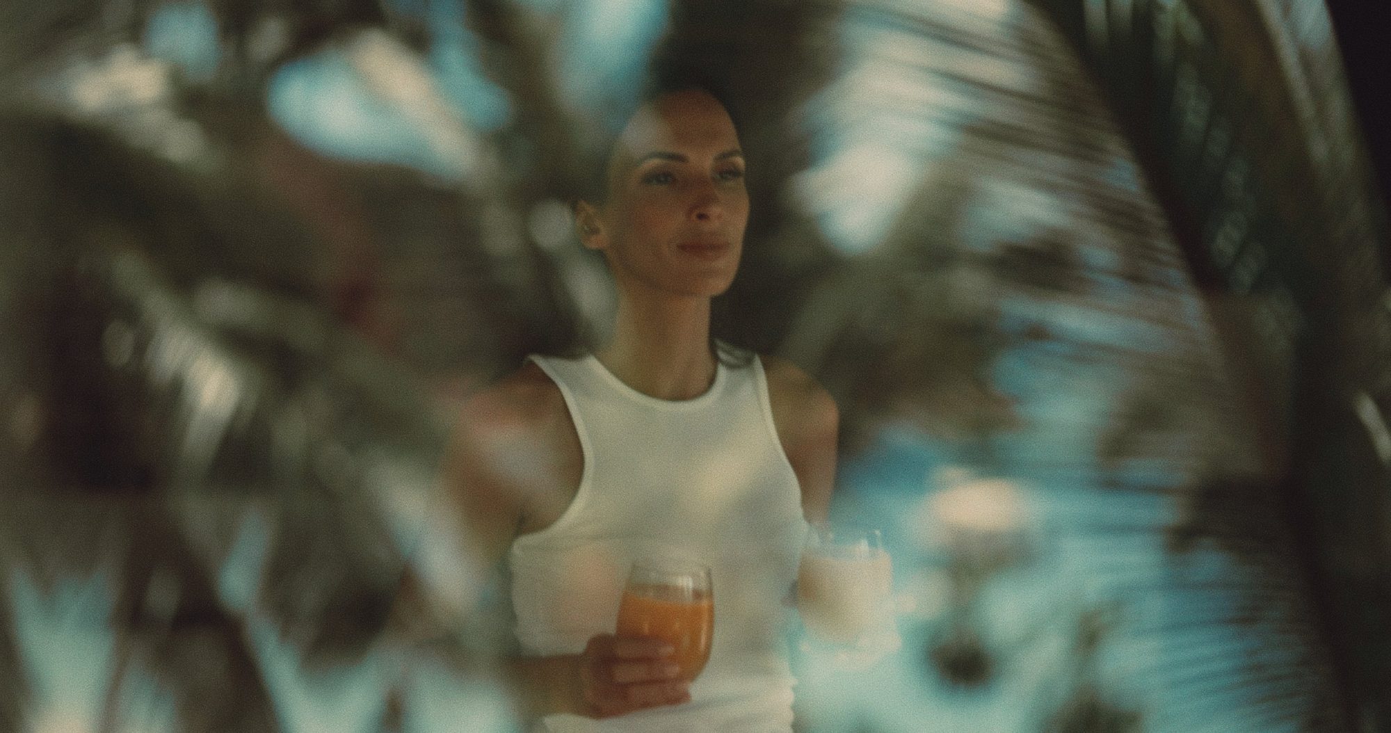 A woman in white carries two drinks, framed through soft palm fronds