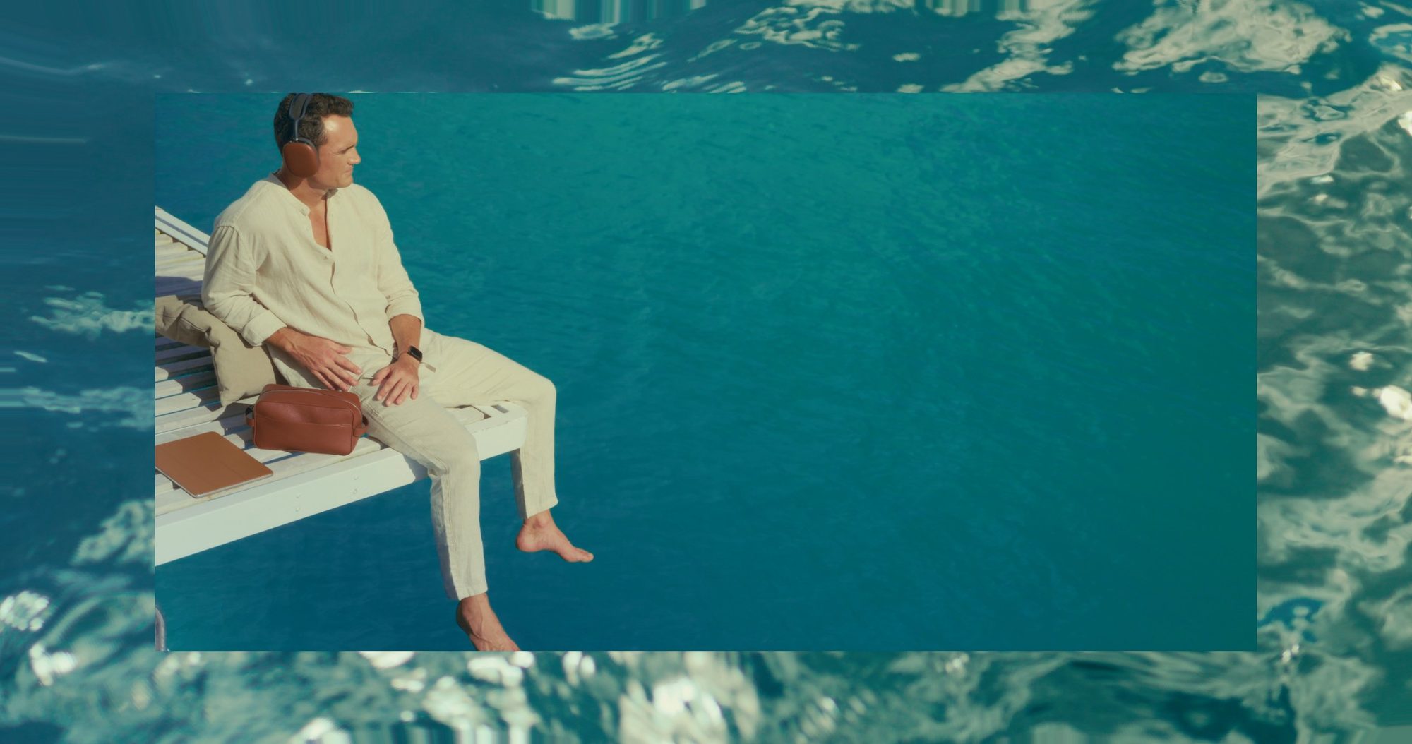 A man in linen with headphones sits on a jetty above blue water