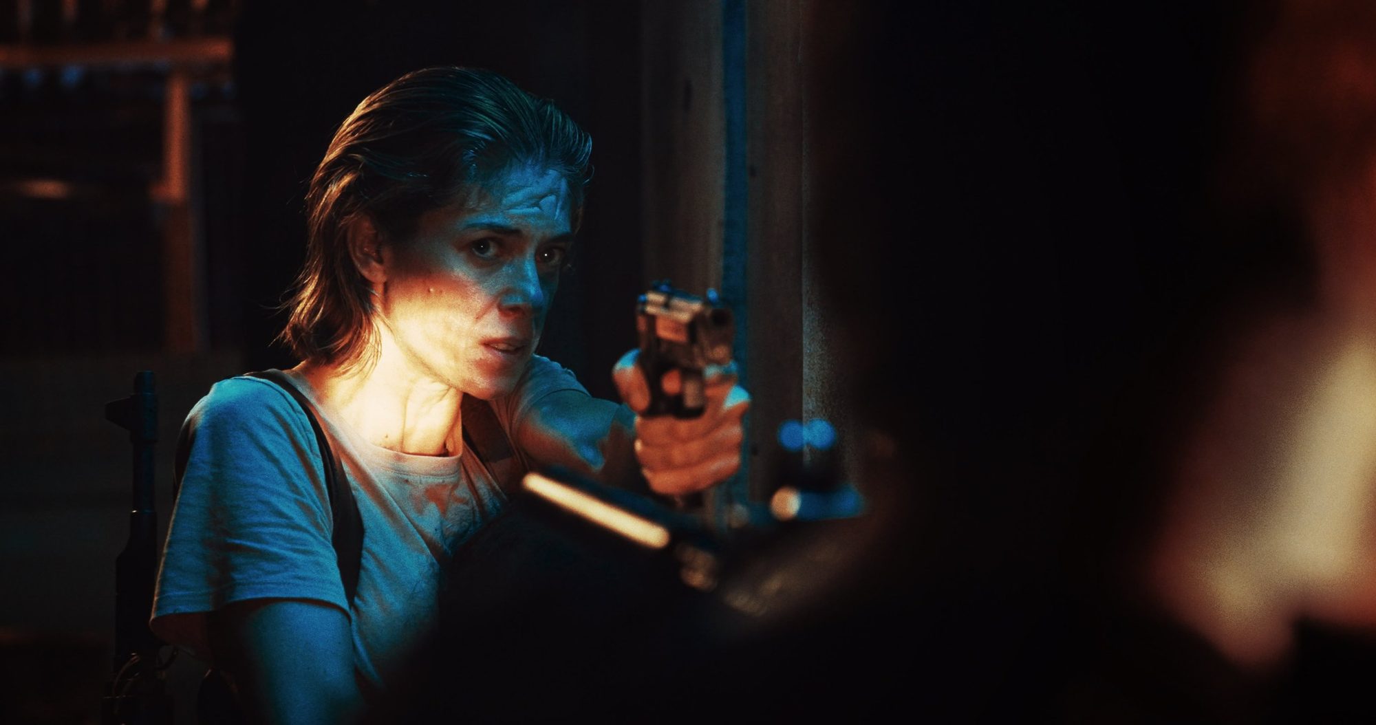 A woman aims a pistol, her face cut by a slash of warm light