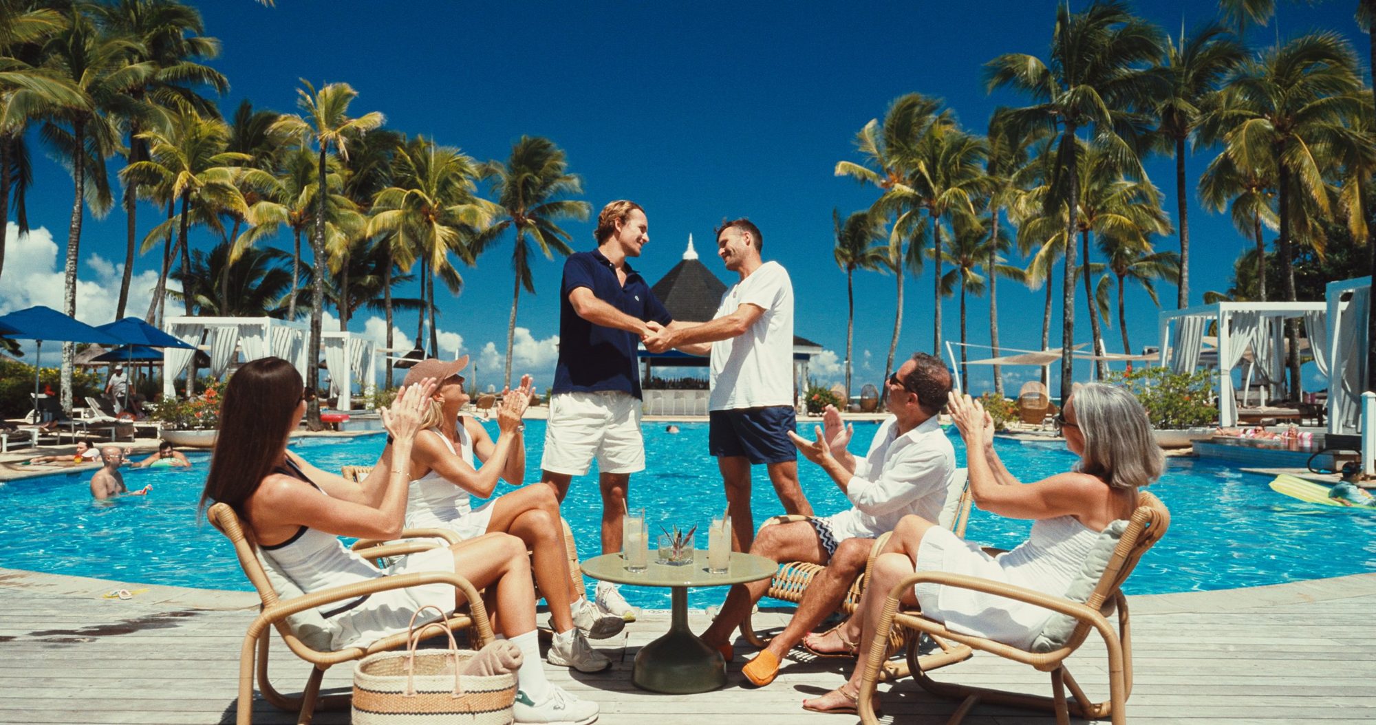 Friends applaud two men shaking hands by a palm-lined pool