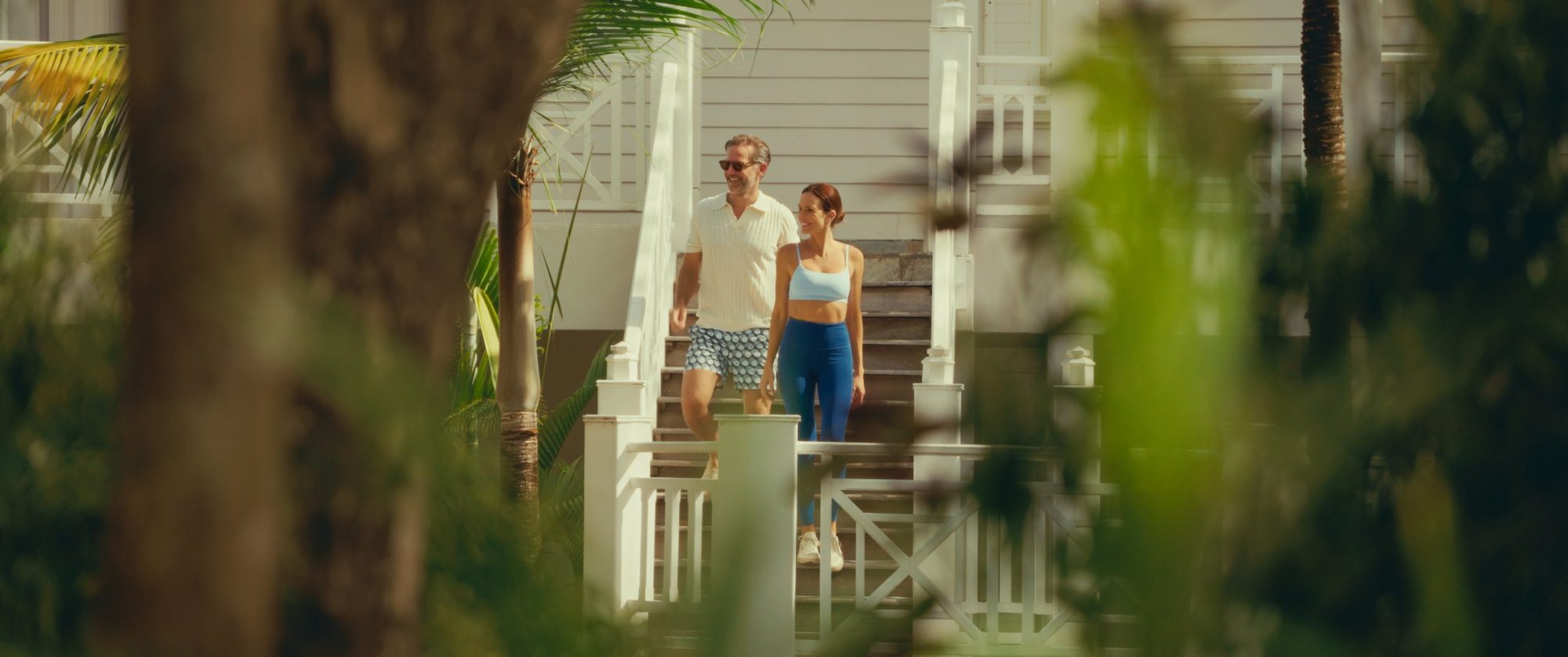 A couple walks down white wooden steps, framed through foliage