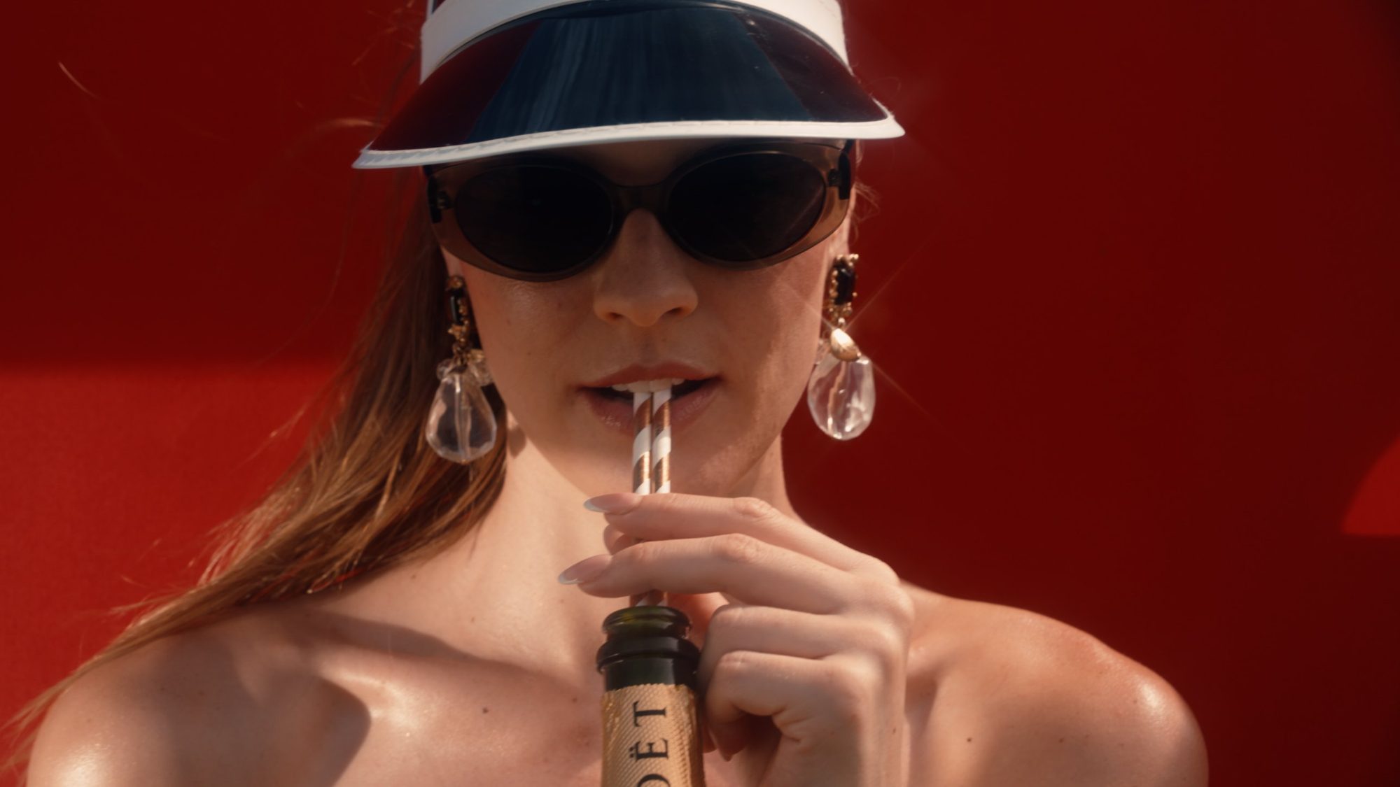 A woman in a visor sips champagne through a straw against a red wall